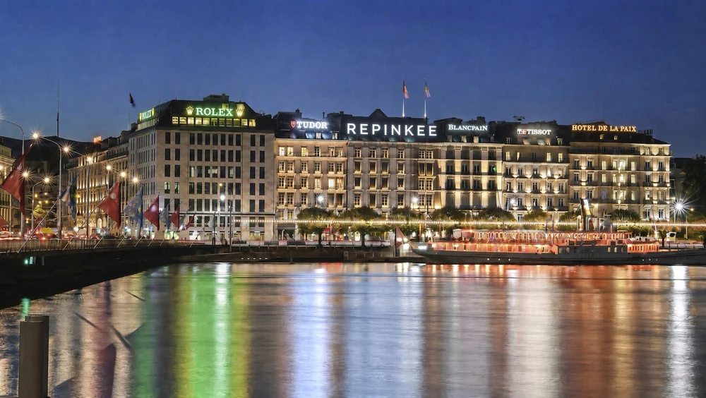 Night view across a lake to a row of lit grand facades, with a Repinkee sign glowing on one of the rooftops
